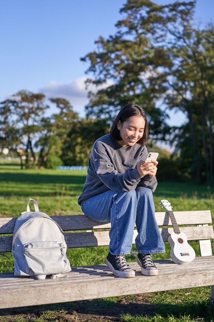 Mujer joven sentada en el parque en un banco con ukelele mirando el mensaje de lectura del teléfono inteligente en el ph móvil