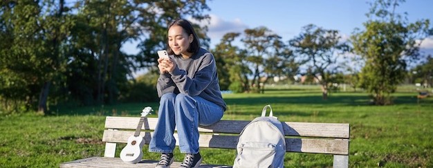 Mujer joven sentada en el parque en un banco con un ukelele mirando un teléfono inteligente leyendo un mensaje en el móvil