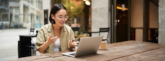 Mujer joven sentada en una reunión en línea en un café al aire libre hablando con la cámara de una computadora portátil explicando algo