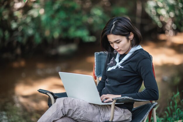 Mujer joven sentada en una silla de camping y usa una computadora portátil mientras se relaja acampando en el espacio de copia del bosque