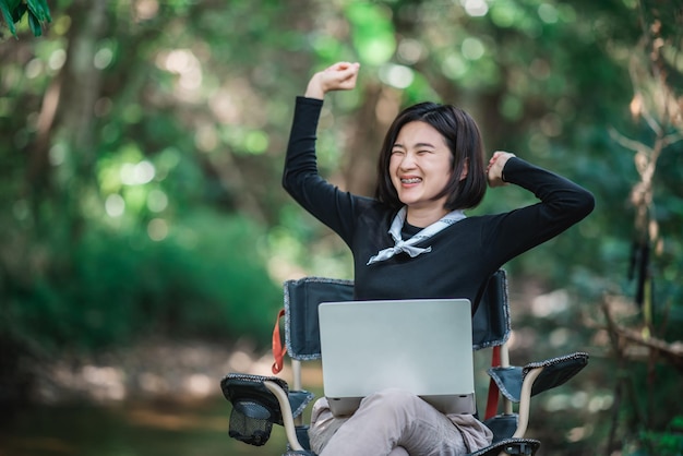 Mujer joven sentada en una silla de camping y usa una computadora portátil mientras se relaja acampando en el espacio de copia del bosque