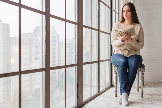 Mujer joven sentada en una silla de madera cerca de la ventana leyendo el libro