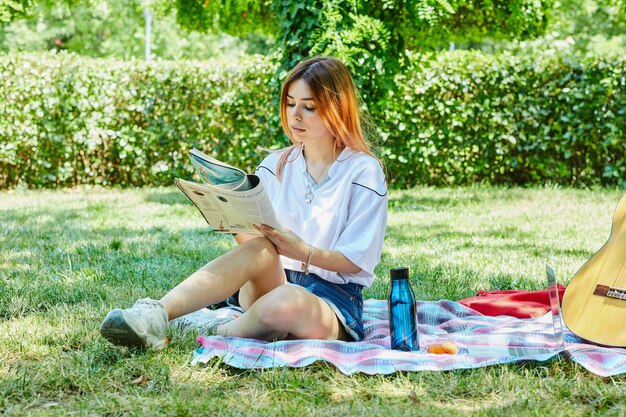 Mujer joven sentada sobre la hierba verde mientras lee el diario junto a la guitarra