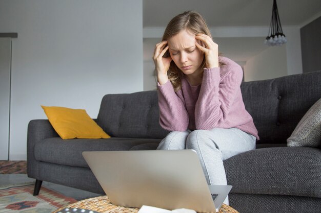 Mujer joven sentada en el sofá con los ojos cerrados, con dolor de cabeza