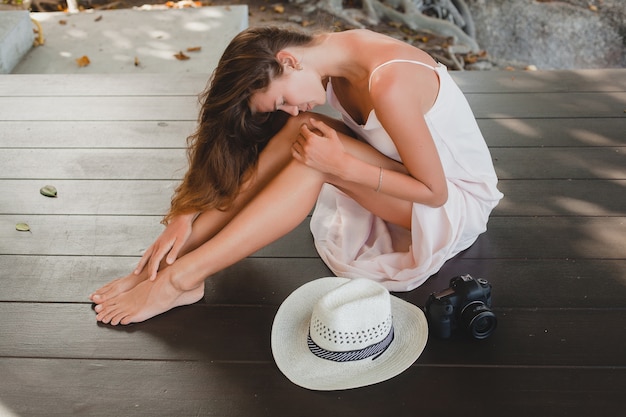 Mujer joven sentada en el suelo descalzo en vestido pálido, sonriente, belleza natural, sombrero de paja, cámara digital,