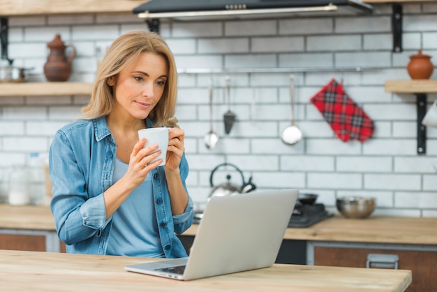 Mujer joven sentada en t él cocina sosteniendo una taza de café mirando portátil