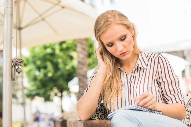 Mujer joven, sentado, en, café al aire libre, escritura, en, cuaderno, con, lápiz