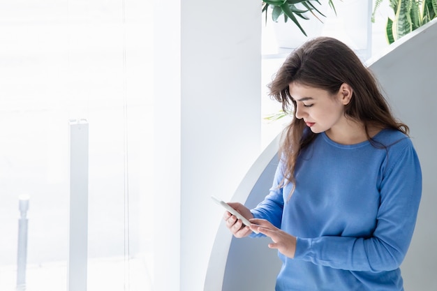 Mujer joven seria atractiva con un suéter azul utiliza un teléfono inteligente en un espacio interior blanco, copia.