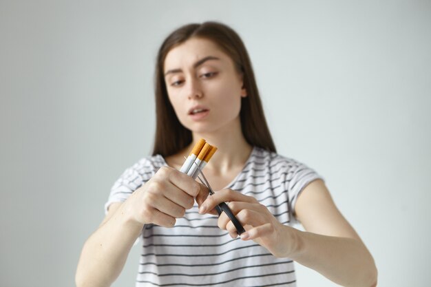 Mujer joven y seria de cabello oscuro con una camiseta a rayas sosteniendo un montón de cigarrillos y tijeras, cortándolos por la mitad mientras decidía dejar de fumar y dejar el mal hábito para siempre. Enfoque selectivo