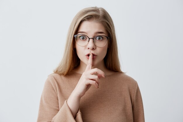 Mujer joven seria con cabello rubio y liso, con lentes, vestida causalmente con un dedo en los labios pidiendo no hacer ruido y ocultar información confidencial privada. Ultra secreto