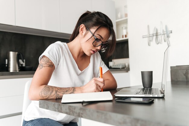 Mujer joven seria concentrada que usa las notas de la escritura de la computadora portátil.