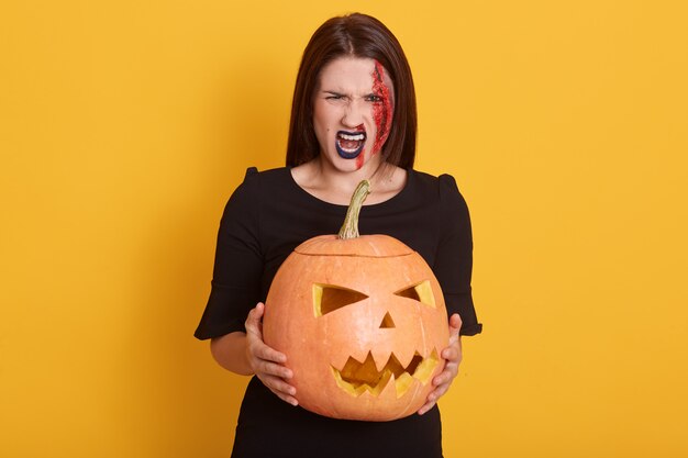 Mujer joven seria con vestido negro, mirando gritando, la señora expresa ira, niña en traje de halloween aislado en amarillo con calabaza en las manos.