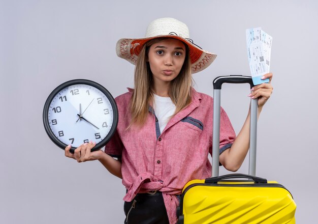 Una mujer joven seria vistiendo camisa roja sosteniendo el reloj de pared maleta amarilla y billetes de avión mientras mira en una pared blanca