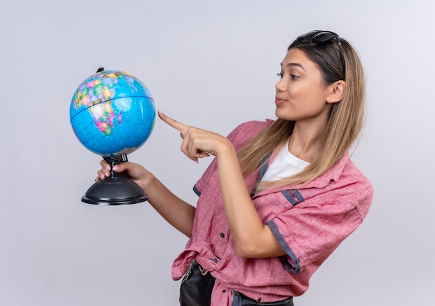 Foto gratuita una mujer joven seria vistiendo la camiseta roja apuntando al mundo con el dedo índice sobre una pared blanca