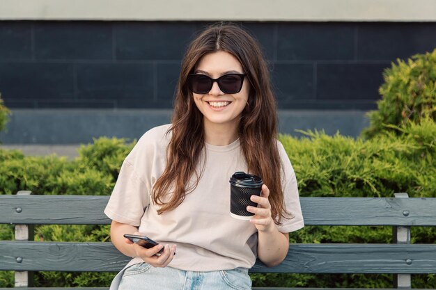 Una mujer joven se sienta en un banco con una taza de café y un teléfono inteligente