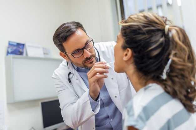 Una mujer joven se sienta en una mesa de examen frente a su médico. El médico se acerca con un depresor de lengua mientras la mujer mira hacia arriba y saca la lengua.