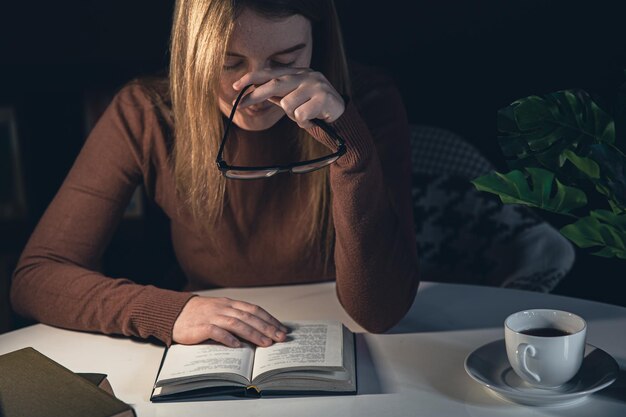 Mujer joven se sienta a la mesa y lee un libro por la noche con una lámpara de luz