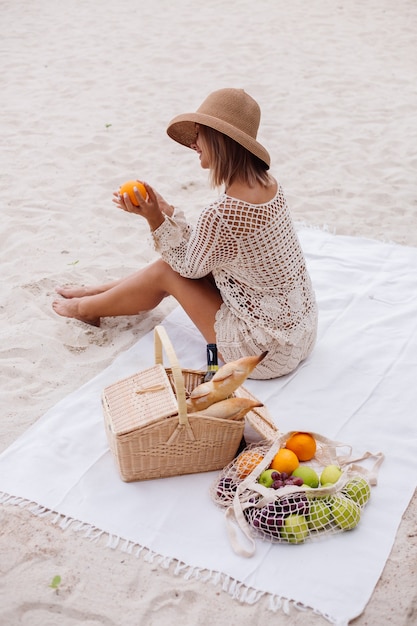 Una mujer joven se sienta en la toalla con un sombrero de paja y una ropa de punto blanca con una cesta de picnic