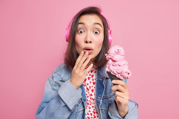 Mujer joven se siente muy sorprendida, no puede creer que sus ojos se queden sin palabras, nota una escena increíble mientras camina por la calle sostiene un gran helado apetitoso escucha música a través de auriculares