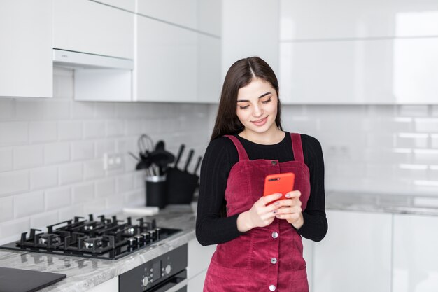 Mujer joven con smartphone apoyado en la mesa de la cocina con taza de café y organizador en un hogar moderno. Mujer sonriente que lee el mensaje de teléfono. Chica morena feliz escribiendo un mensaje de texto