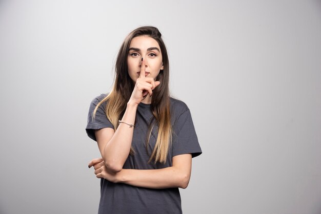 Mujer joven sobre un fondo gris que muestra un gesto de silencio poniendo el dedo en la boca.