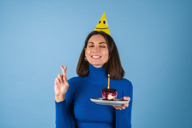 Mujer joven sobre una pared azul celebra un cumpleaños, sostiene un trozo de tarta, feliz, emocionado, pide un deseo