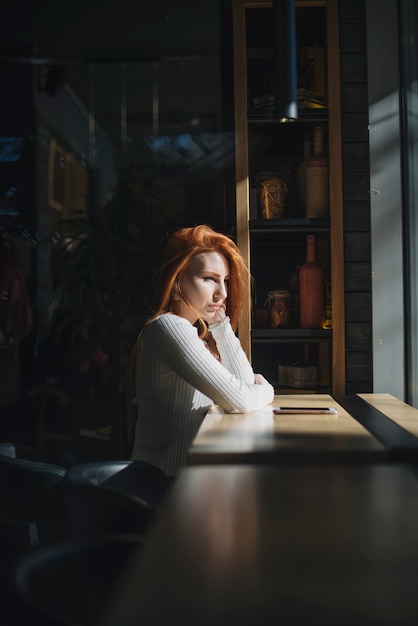 Una mujer joven solitaria que se sienta cerca de la ventana con el teléfono móvil en la tabla