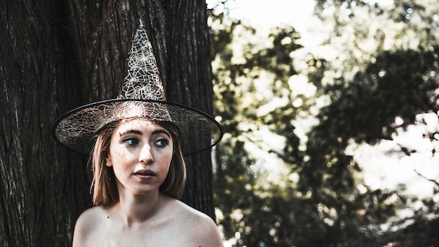 Mujer joven en sombrero de bruja de pie cerca de árbol