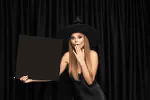 Foto gratuita mujer joven con sombrero como bruja sosteniendo tablero negro contra cortinas negras