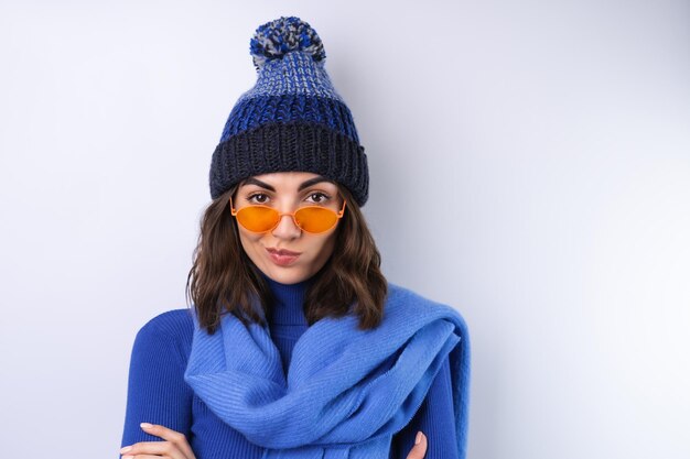 Mujer joven en un sombrero de cuello alto de golf azul y bufanda gafas de sol sobre un fondo blanco.