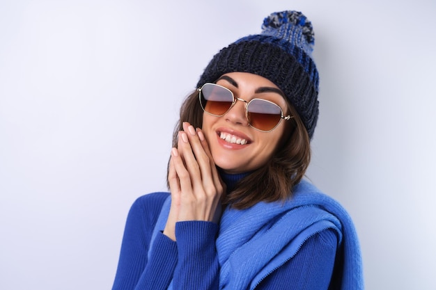 Mujer joven en un sombrero de cuello alto de golf azul y gafas de sol de bufanda sobre un fondo blanco alegre de buen humor