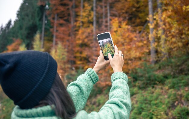 Una mujer joven con un sombrero negro toma una foto del bosque de otoño en un teléfono inteligente