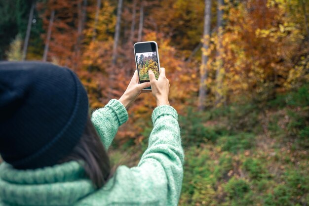 Una mujer joven con un sombrero negro toma una foto del bosque de otoño en un teléfono inteligente