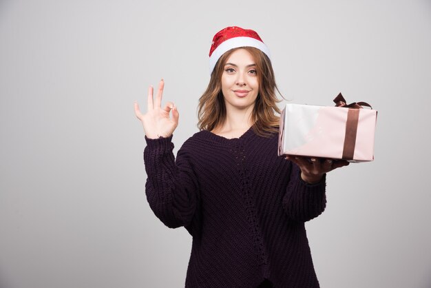 Mujer joven con sombrero de Papá Noel con un presente que muestra un gesto bien.