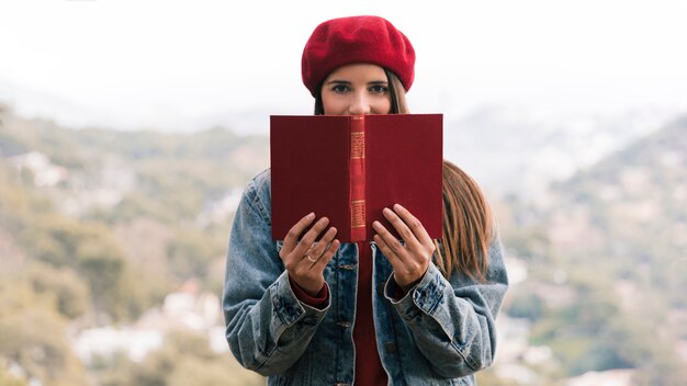 Mujer joven con sombrero de punto sobre su cabeza sosteniendo un libro frente a su boca