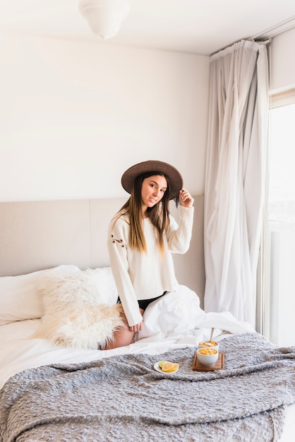 Foto gratuita mujer joven con sombrero de rodillas en la cama con el desayuno en el dormitorio