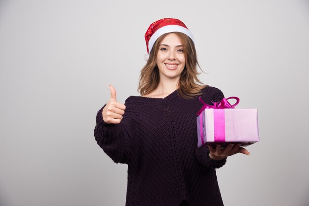 Mujer joven con sombrero de Santa con caja de regalo mostrando un pulgar hacia arriba.