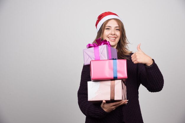 Mujer joven con sombrero de Santa con cajas de regalo mostrando un pulgar hacia arriba.
