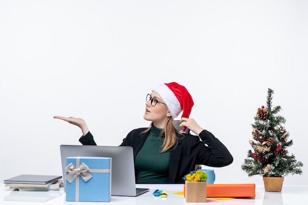 Mujer joven con sombrero de santa claus y anteojos sentado en una mesa con un árbol de Navidad y un regalo