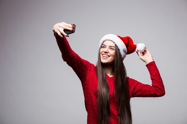 Mujer joven con sombrero de santa tomar selfie en el teléfono aislado en la pared gris