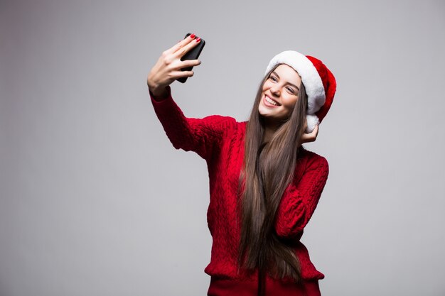 Mujer joven con sombrero de santa tomar selfie en el teléfono aislado en la pared gris