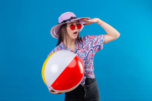 Mujer joven con sombrero de verano con gafas de sol rojas con bola inflable mirando lejos con la mano buscando a alguien sobre la pared azul