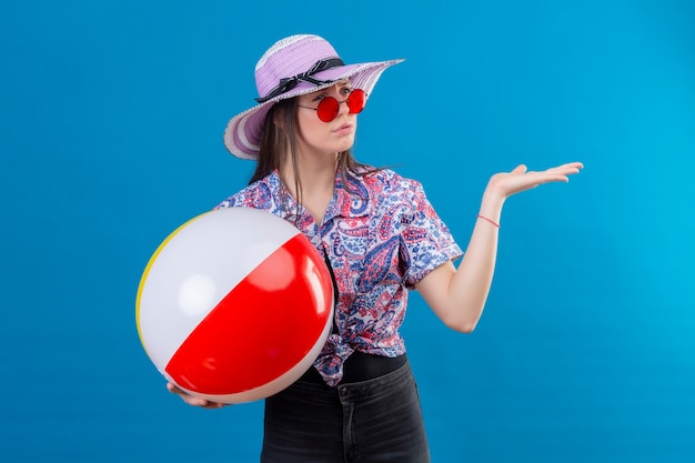 Mujer joven con sombrero de verano con gafas de sol rojas sosteniendo una pelota inflable mirando a un lado con el ceño fruncido de pie disgustado con el brazo levantado sobre fondo azul