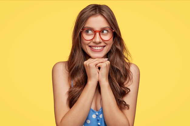 Mujer joven soñadora con gafas posando contra la pared amarilla
