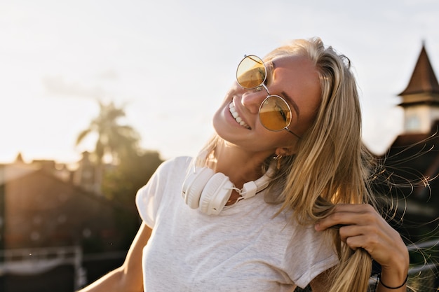 Mujer joven soñadora lleva grandes auriculares blancos posando sobre fondo de cielo con linda sonrisa.