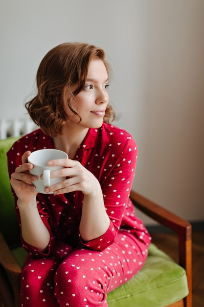 Mujer joven soñadora en pijama rojo sosteniendo una taza de café. Filmación en interiores de mujer pensativa sentada en un sillón y mirando a otro lado.