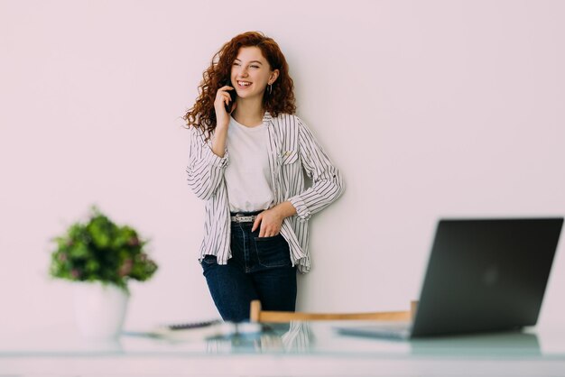 Mujer joven soñadora sentada detrás de una computadora portátil moderna usando un teléfono moderno trabajando en casa y hablando con el cliente sobre el concepto de su proyecto