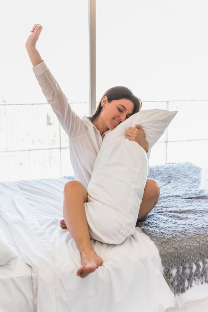 Mujer joven soñolienta que sostiene la almohada blanca que se sienta en la cama que estira sus manos