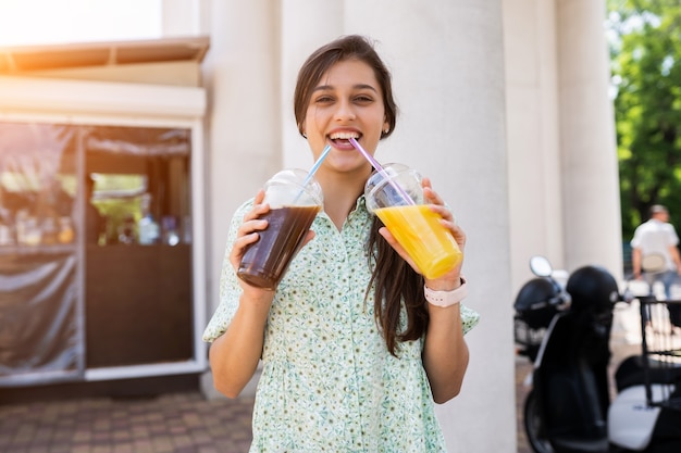 Mujer joven sonriendo y bebe dos cócteles con hielo en vasos de plástico con paja en las calles de la ciudad.