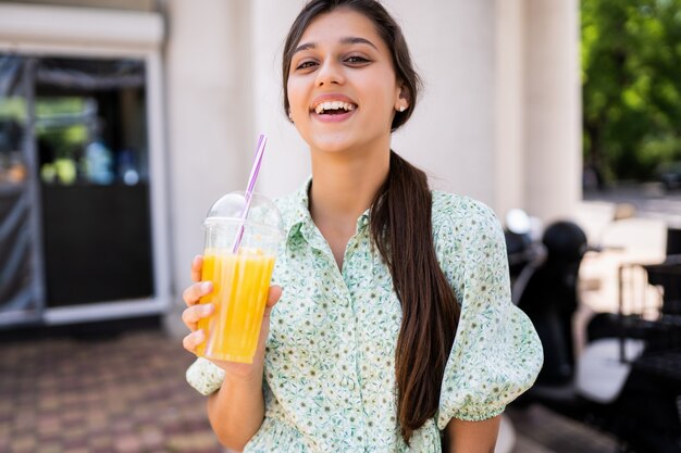 Mujer joven sonriendo y bebiendo cócteles con hielo en vaso de plástico con paja en las calles de la ciudad.